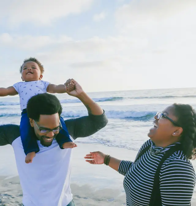 A father lifts his laughing baby onto his shoulders on a sunny beach while the mother smiles beside them