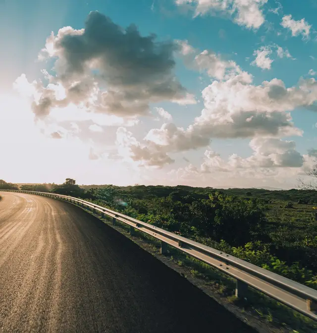 An empty tropical road curving past green coastal scrub toward a low sun