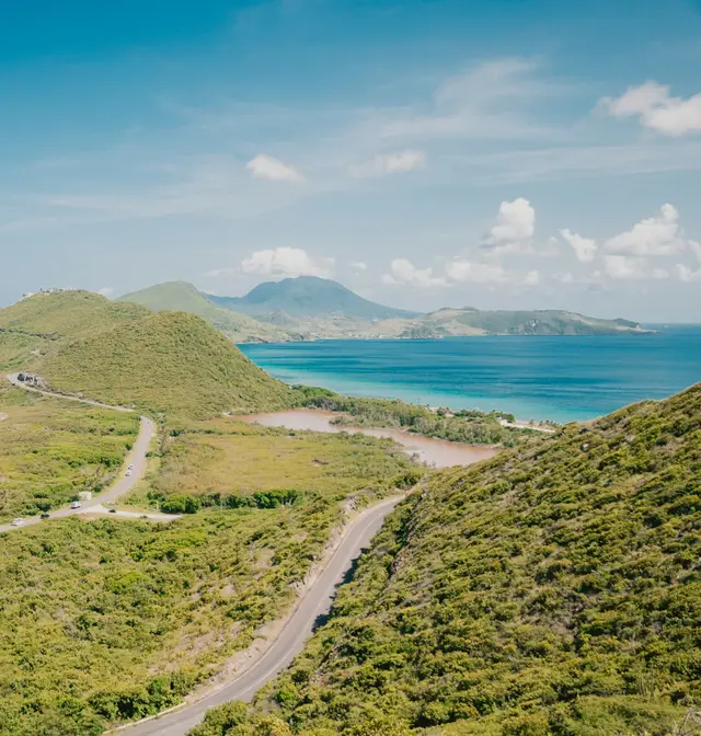A road winding along green hills above turquoise bays on the south-east coast of St Kitts, with Nevis on the horizon