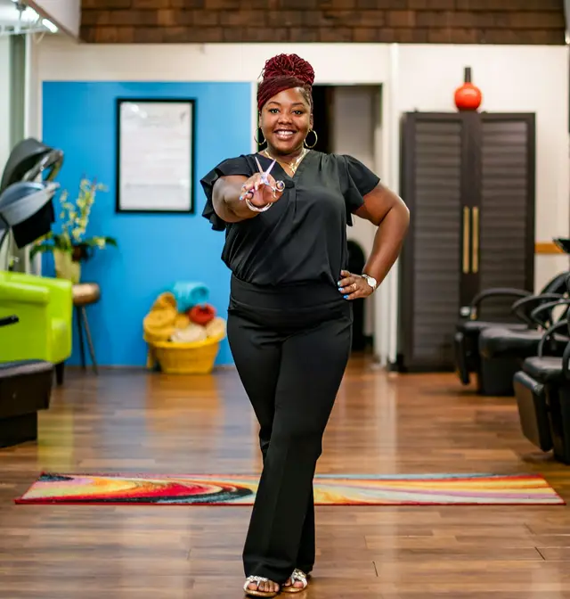 A smiling salon owner in a red headwrap standing in her bright hair salon
